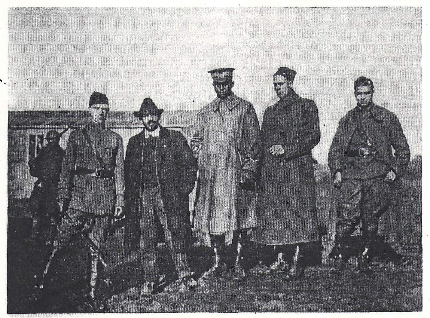 A line of five men in old-fashioned military clothing and hats look seriously at the camera. Some of them wear long coats and boots. The men on the far left and right are wearing military uniforms. Another man stands behind the group, with his back to the camera, in front of a low, rectangular building. The image is black and white.