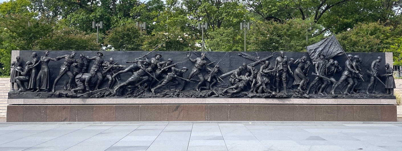 A 58-foot wide bronze wall sculpture with carvings of 38 figures that represent an American soldier's World War One war experience. It sits atop a stone base in an open area, backed by leafy green trees.
