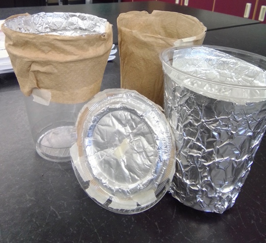 Student prototype projects made from plastic cups. Three clear plastic cups, some wrapped in foil and others wrapped in brown paper towels or aluminum foil, sit together on a grey table. A clear plastic cup lid, with foil taped to the top, leans against the cups.